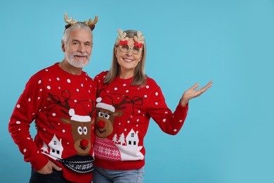 Photo of Senior couple in Christmas sweaters, reindeer headband and party glasses on light blue background. Space for text