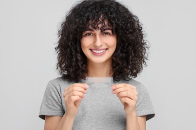 Young woman holding teeth whitening strips on light grey background