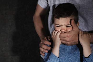 Adult man covering scared little boy's mouth on dark background, space for text. Child in danger