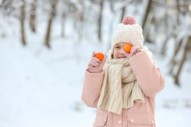 Photo of Cute little girl covering eye with tangerine in snowy park on winter day, space for text