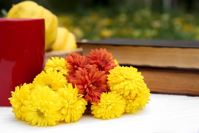 Photo of Beautiful chrysanthemum flowers, cup and books on white wooden table outdoors, closeup