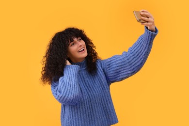 Beautiful young woman taking selfie with smartphone on orange background