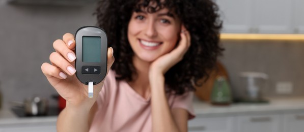 Photo of Diabetes. Happy woman holding digital glucometer in kitchen, selective focus