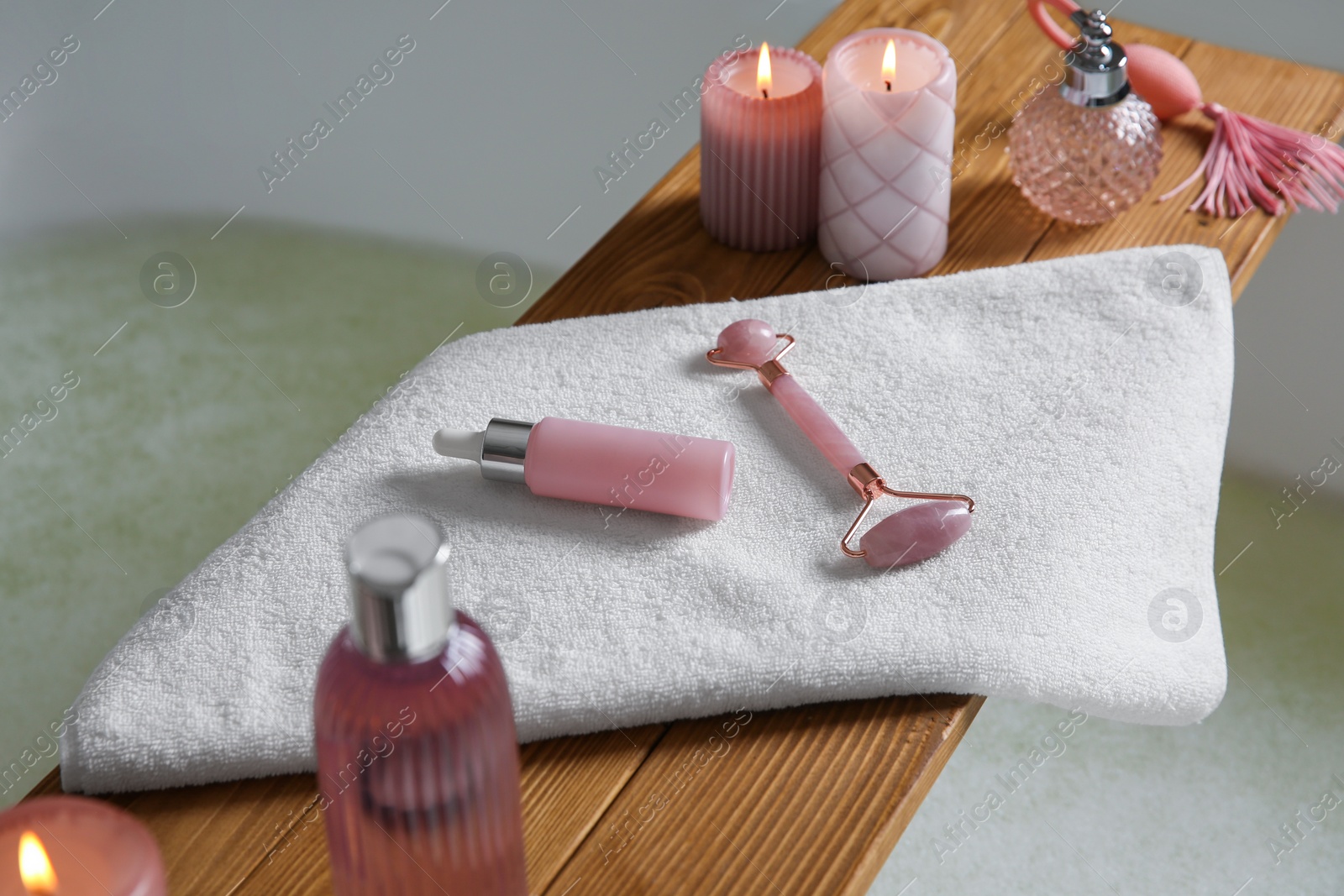 Photo of Wooden bath tray with face roller, cosmetic products and candles on tub indoors