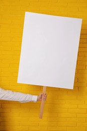 Woman holding blank sign near yellow brick wall, closeup. Space for design