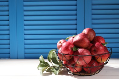 Metal bowl with wet red apples and green leaves on white table. Space for text