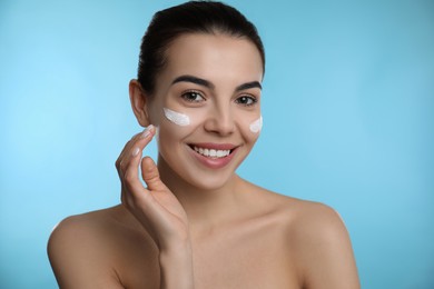 Young woman applying facial cream on light blue background