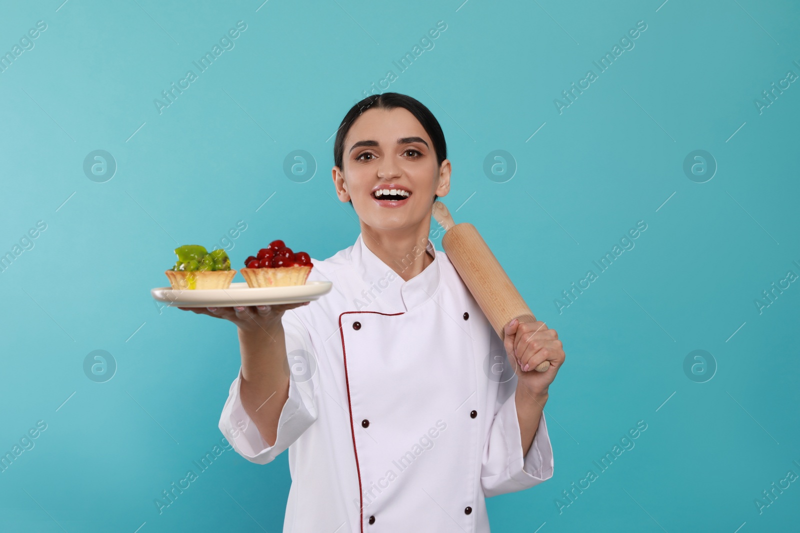 Photo of Happy professional confectioner in uniform with delicious tartlets and rolling pin on light blue background