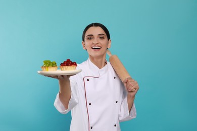 Happy professional confectioner in uniform with delicious tartlets and rolling pin on light blue background