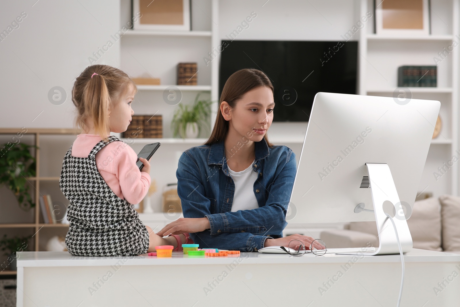 Photo of Woman working remotely at home. Woman using computer while her daughter playing with phone. Child sitting on desk