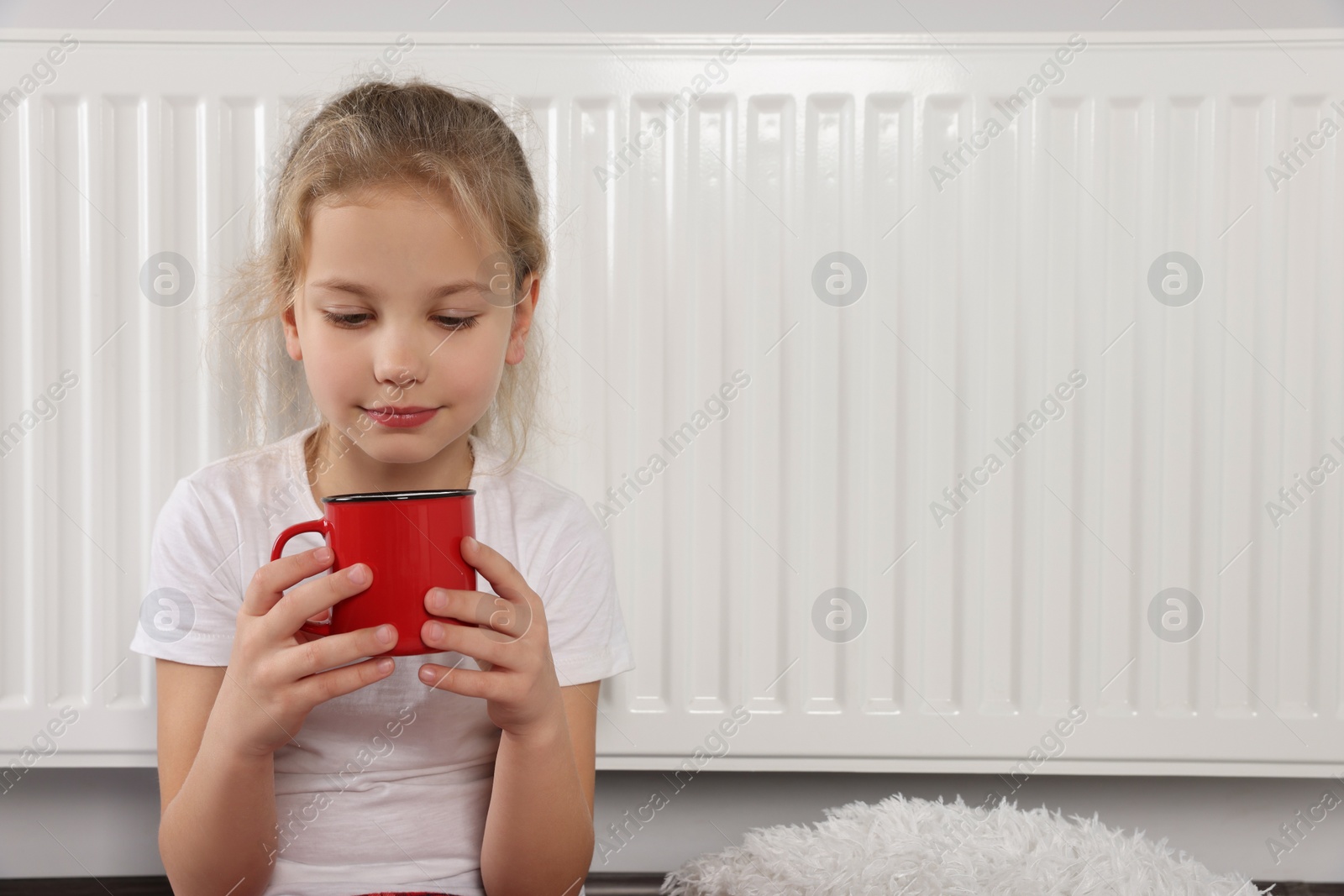 Photo of Little girl with cup of drink near heating radiator indoors, space for text