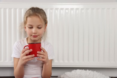 Little girl with cup of drink near heating radiator indoors, space for text