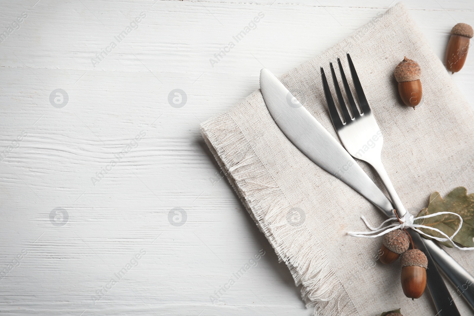 Photo of Cutlery, acorns and napkin on white wooden background, flat lay with space for text. Table setting elements