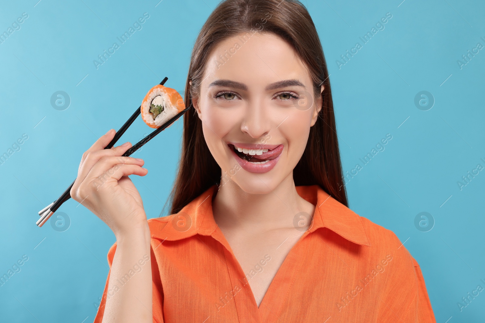 Photo of Happy young woman holding sushi roll with chopsticks on light blue background