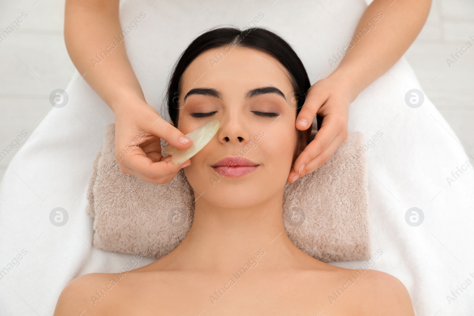 Photo of Young woman receiving facial massage with gua sha tool in beauty salon, top view