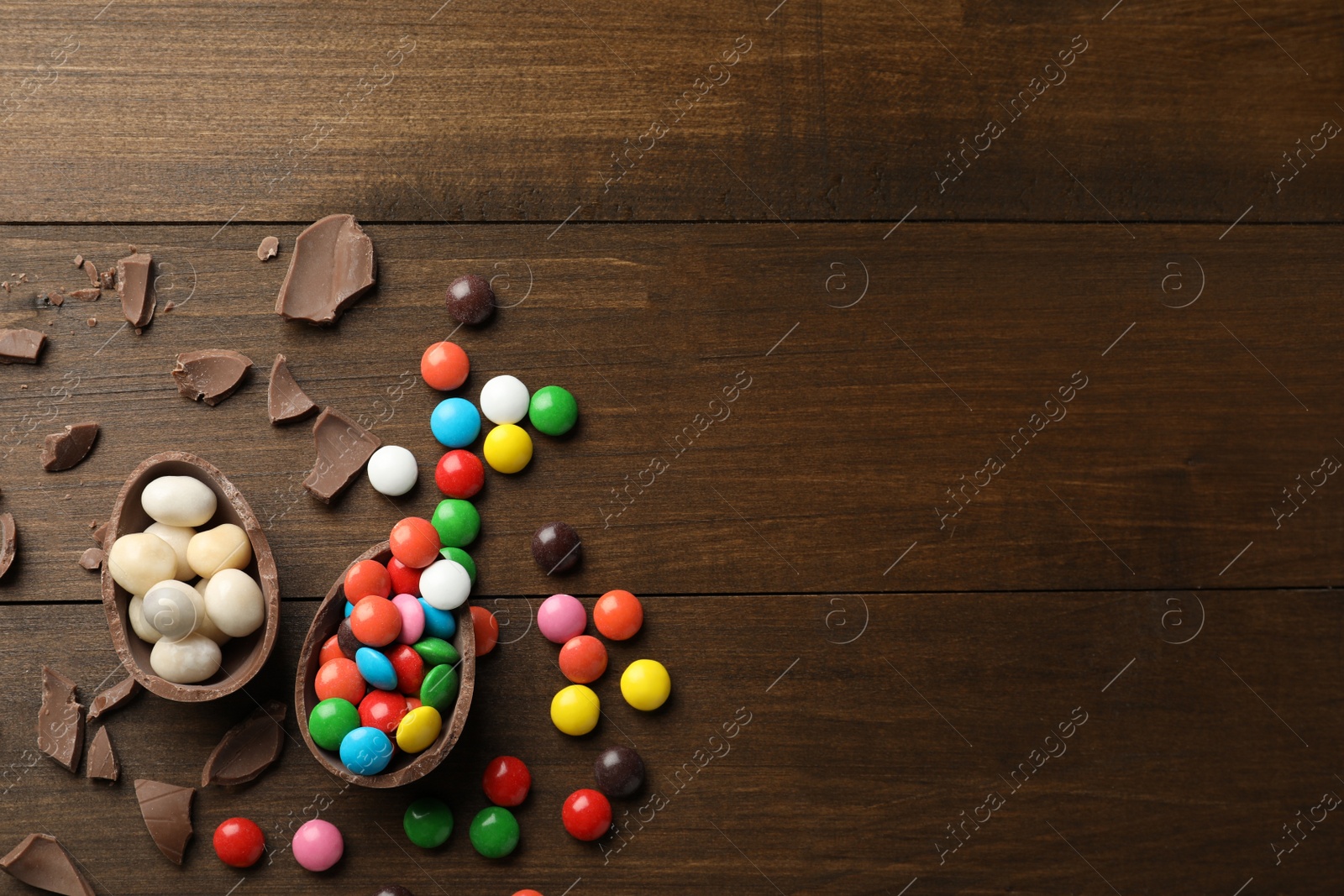 Photo of Broken chocolate egg and colorful candies on wooden table, flat lay. Space for text