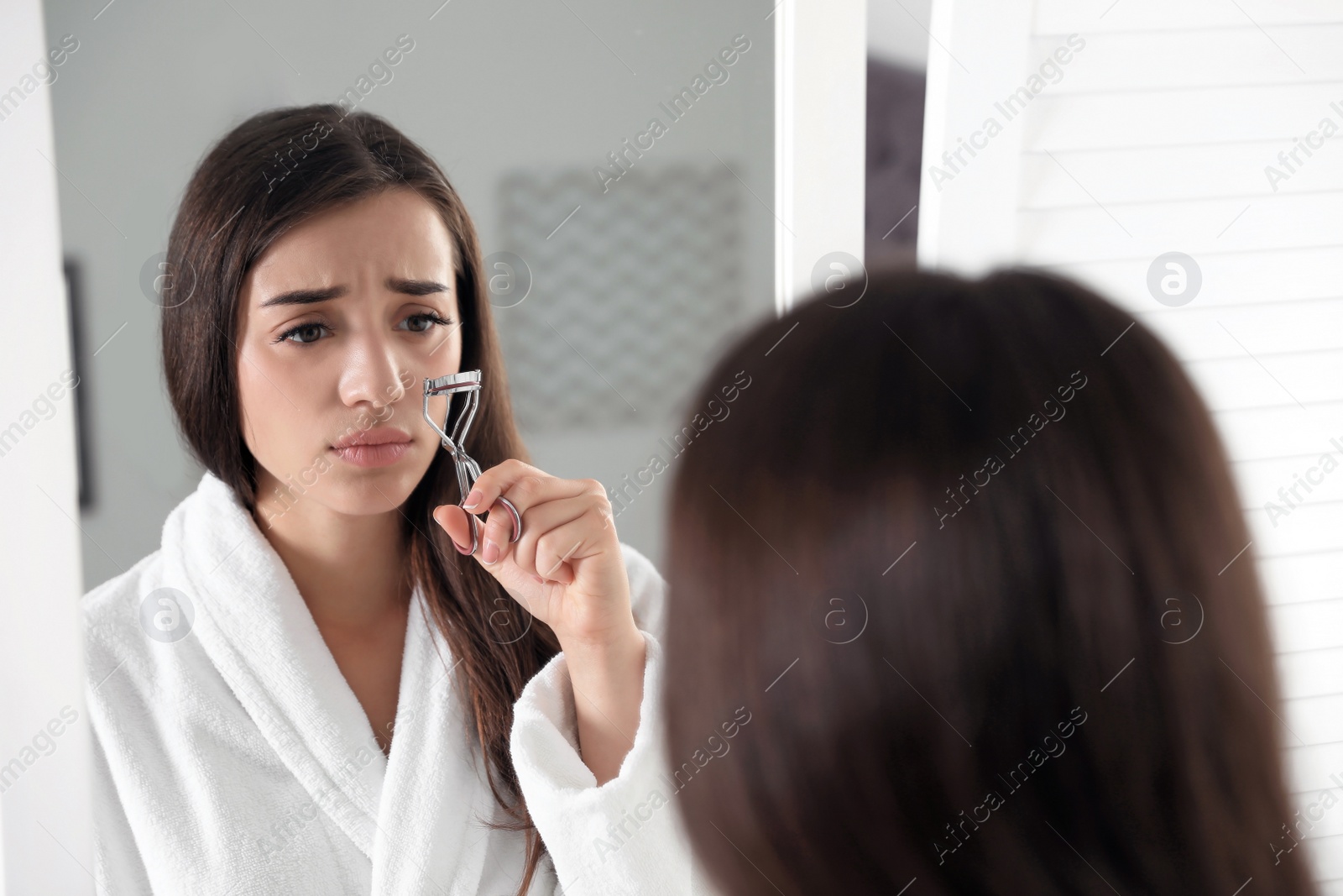 Photo of Emotional young woman curling her eyelashes near mirror indoors