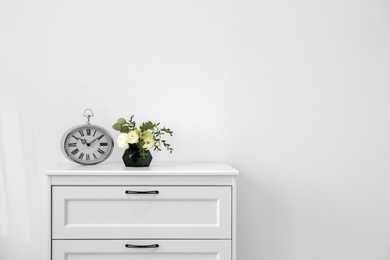 Modern chest of drawers with fresh flowers and clock near wall. Space for text