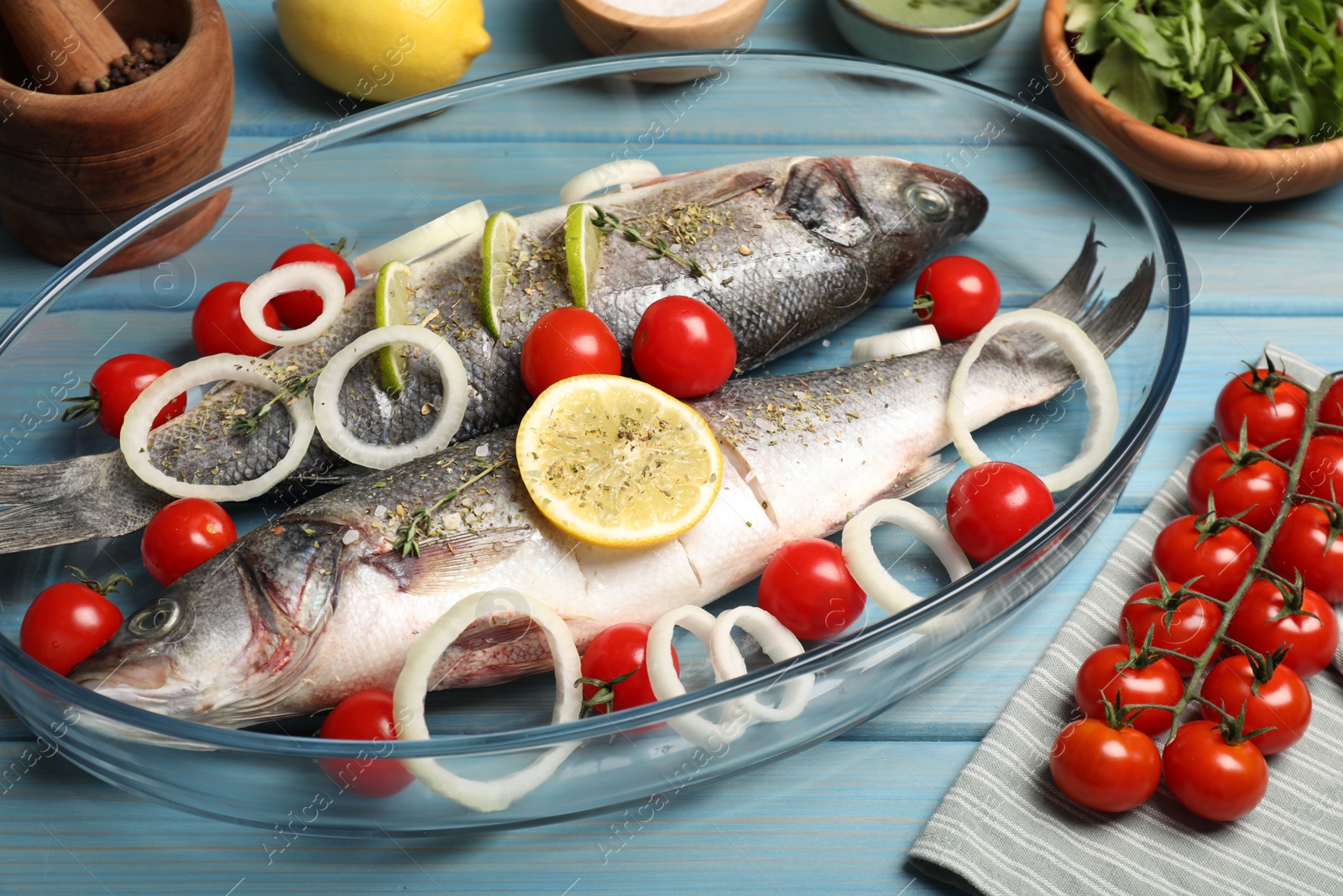 Photo of Glass baking tray with sea bass fish and ingredients on light blue wooden table, closeup