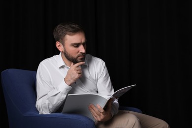 Photo of Handsome man reading magazine in armchair against dark background