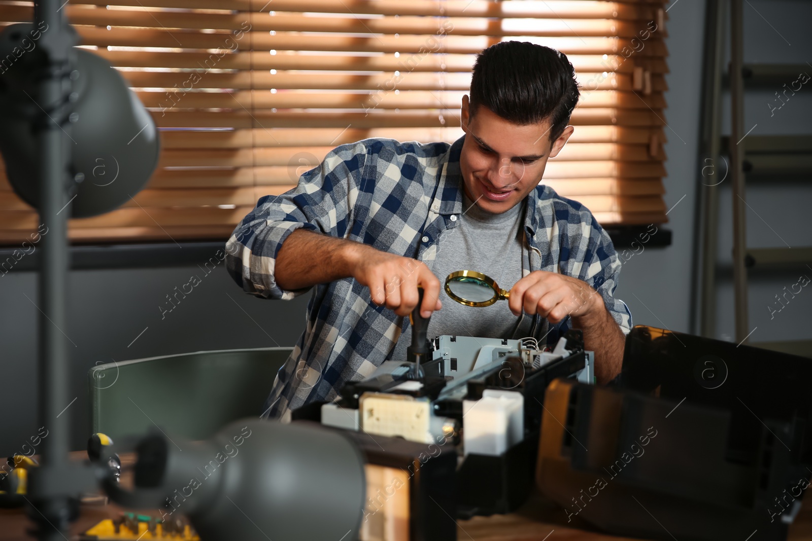 Photo of Repairman with magnifying glass and screwdriver fixing modern printer in office