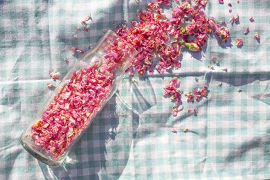 Glass bottle with dry tea rose petals on checkered fabric, top view