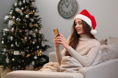 Beautiful young smiling woman in Santa hat using smartphone near Christmas tree at home, space for text