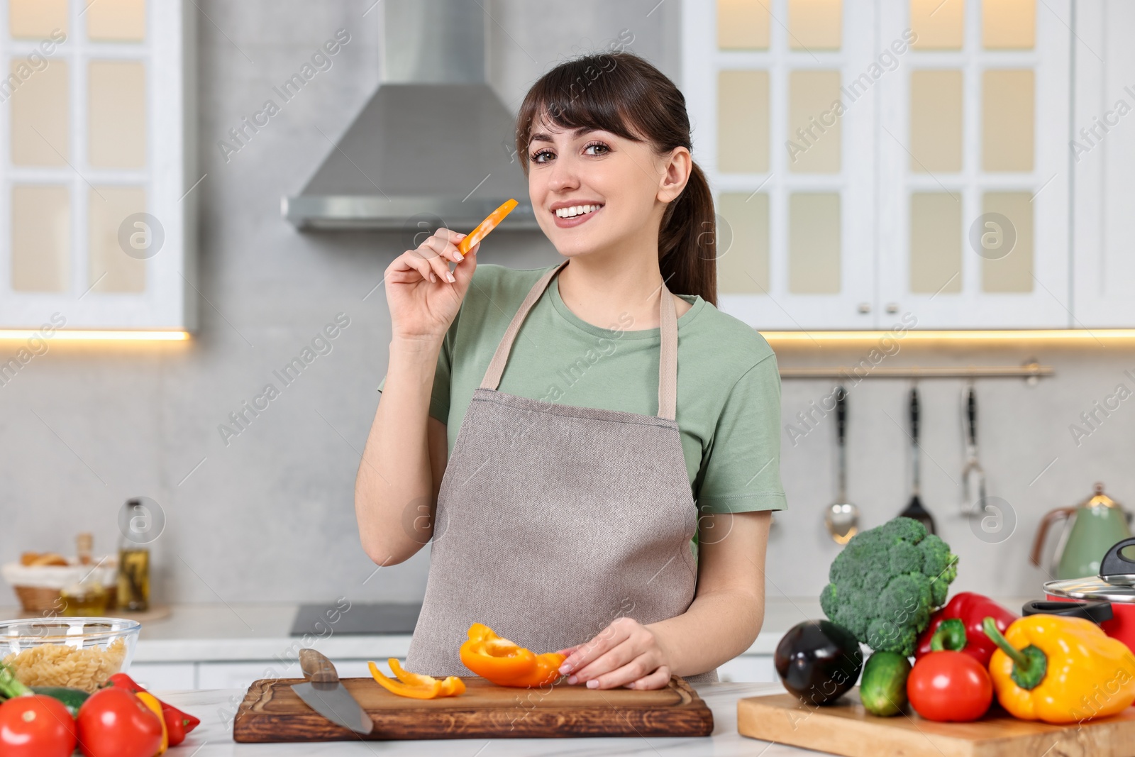 Photo of Happy young housewife with cut bell pepper cooking at white marble table in kitchen