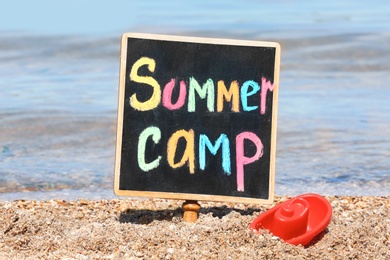 Photo of Little blackboard with text SUMMER CAMP and toy on sand near sea