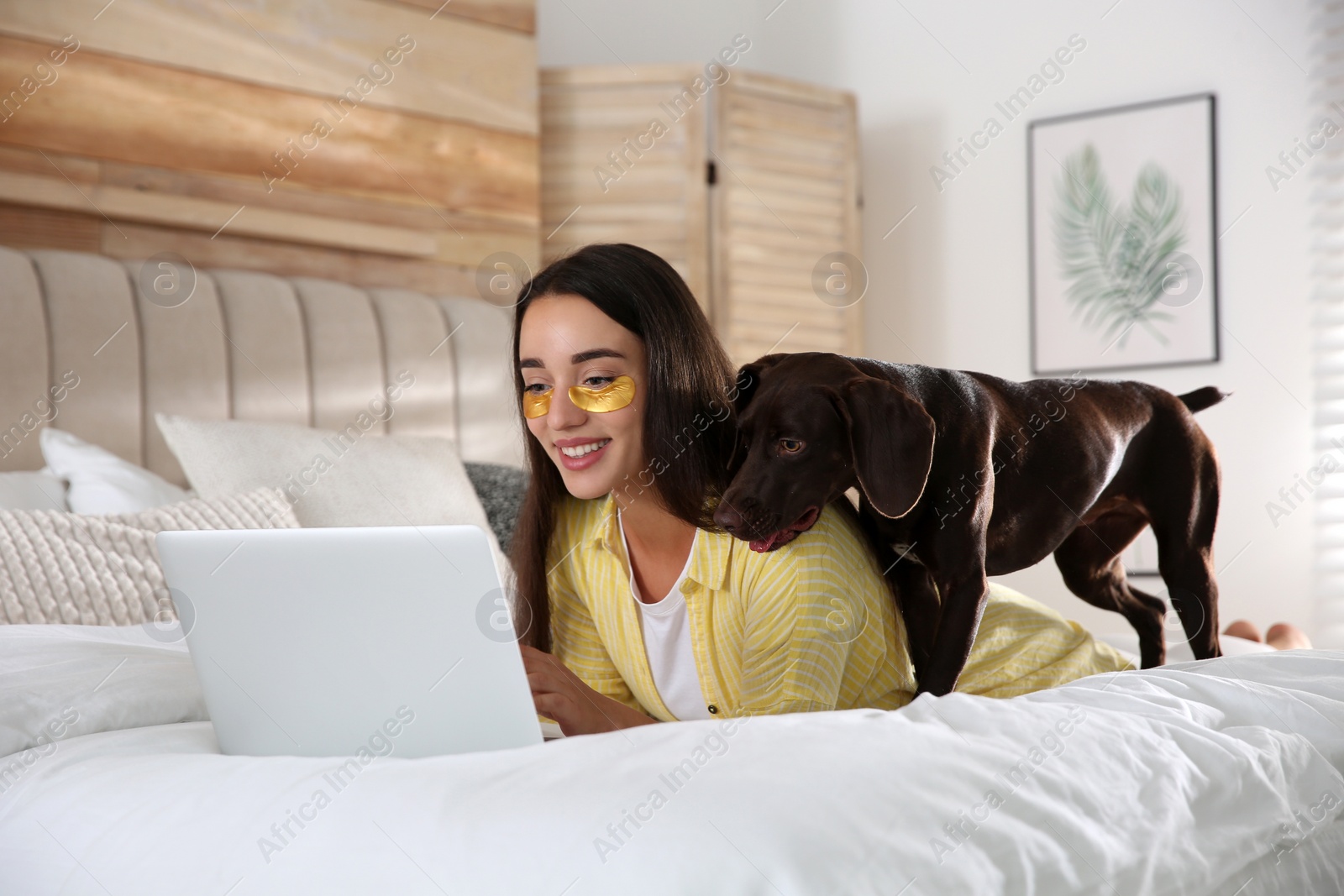 Photo of Young woman with eye patches working on laptop near her dog in bedroom. Home office concept