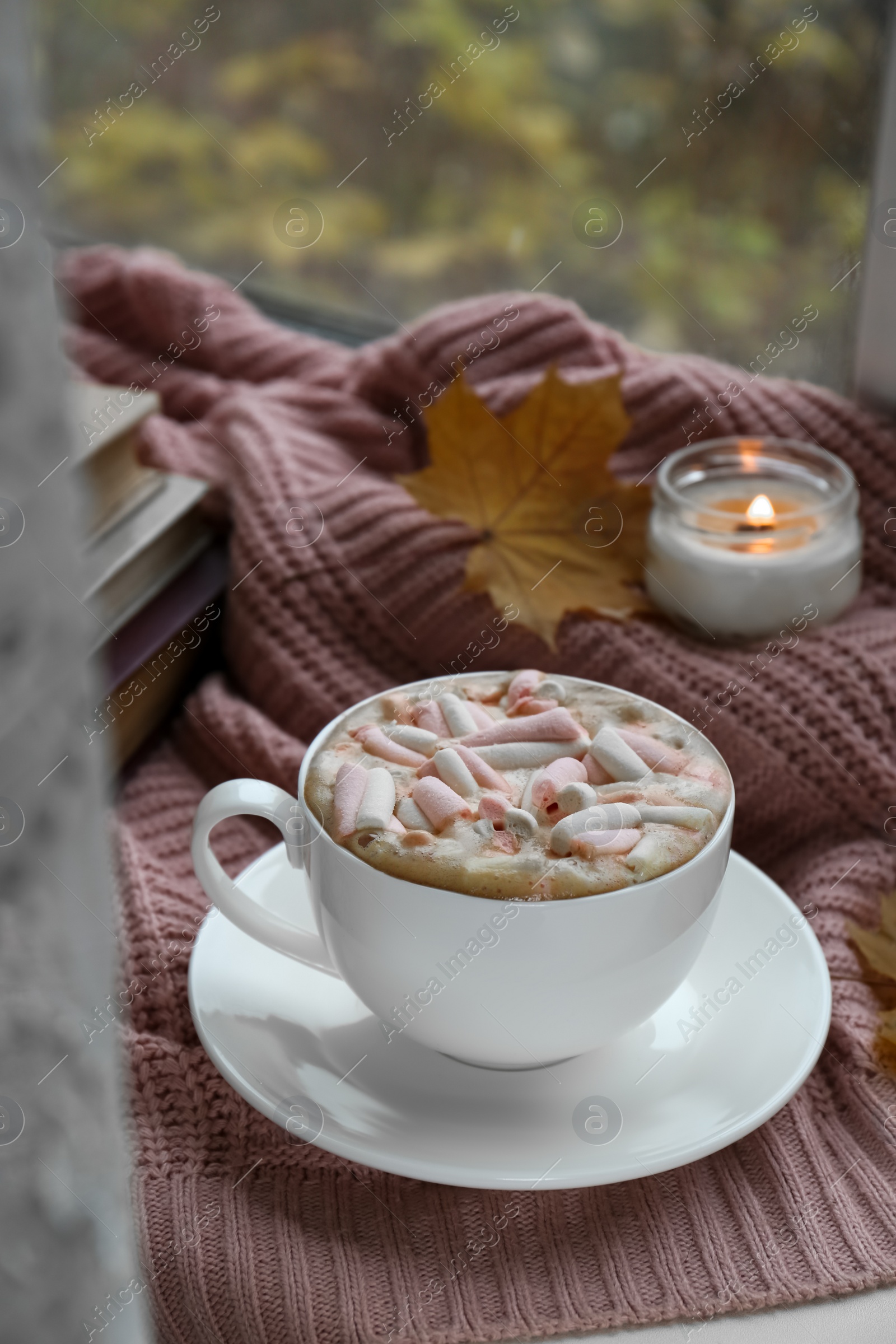 Photo of Cup of tasty hot drink with marshmallows, books, leaf and burning candle on blanket. Autumn coziness