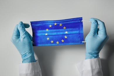 Doctor holding medical mask with European Union flag on light background, closeup. Coronavirus outbreak in Europe