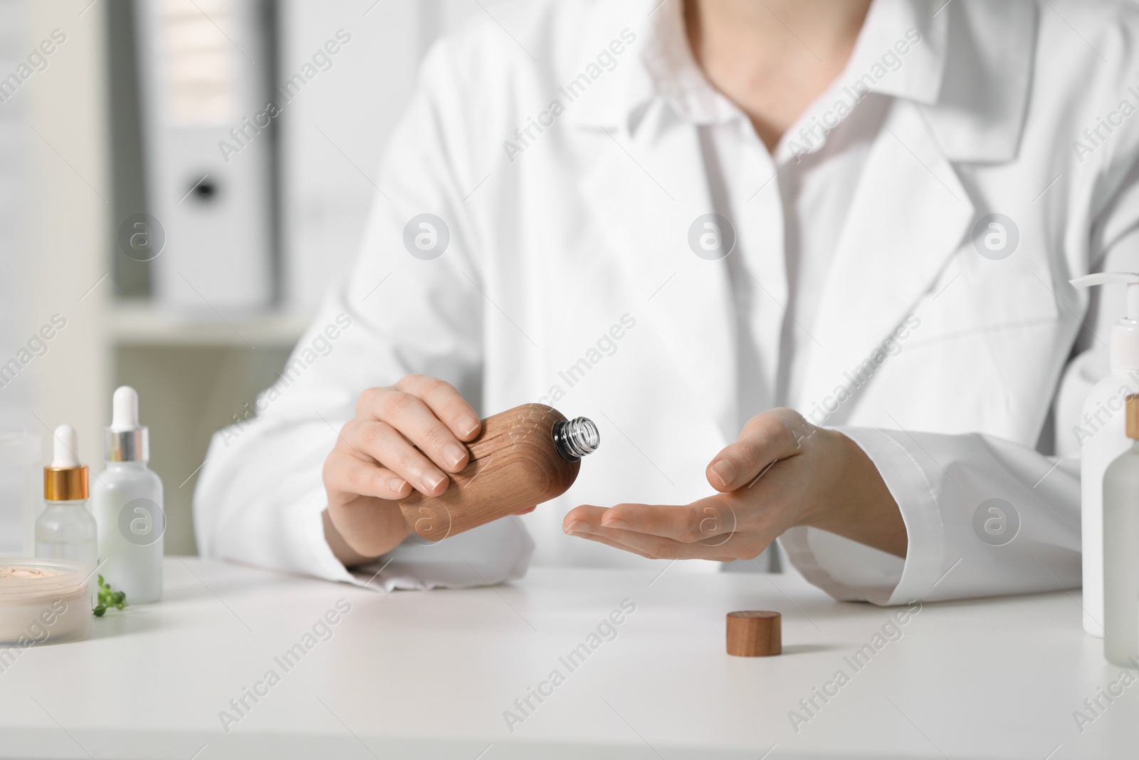 Photo of Dermatologist with bottle testing cosmetic product at white table indoors, selective focus