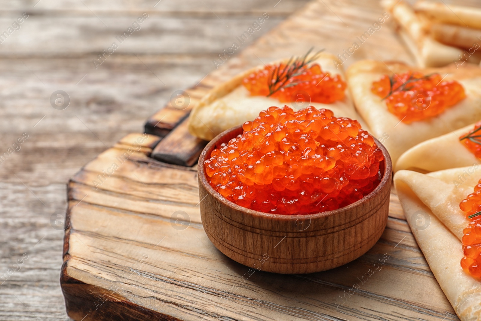 Photo of Bowl with delicious red caviar and thin pancakes on wooden board