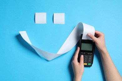 Woman using payment terminal and smiley face made of thermal paper for receipt on light blue background, top view