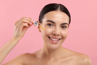 Beautiful young woman applying serum onto her face on pink background