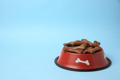 Photo of Red bowl with bone shaped dog cookies on light blue background, space for text