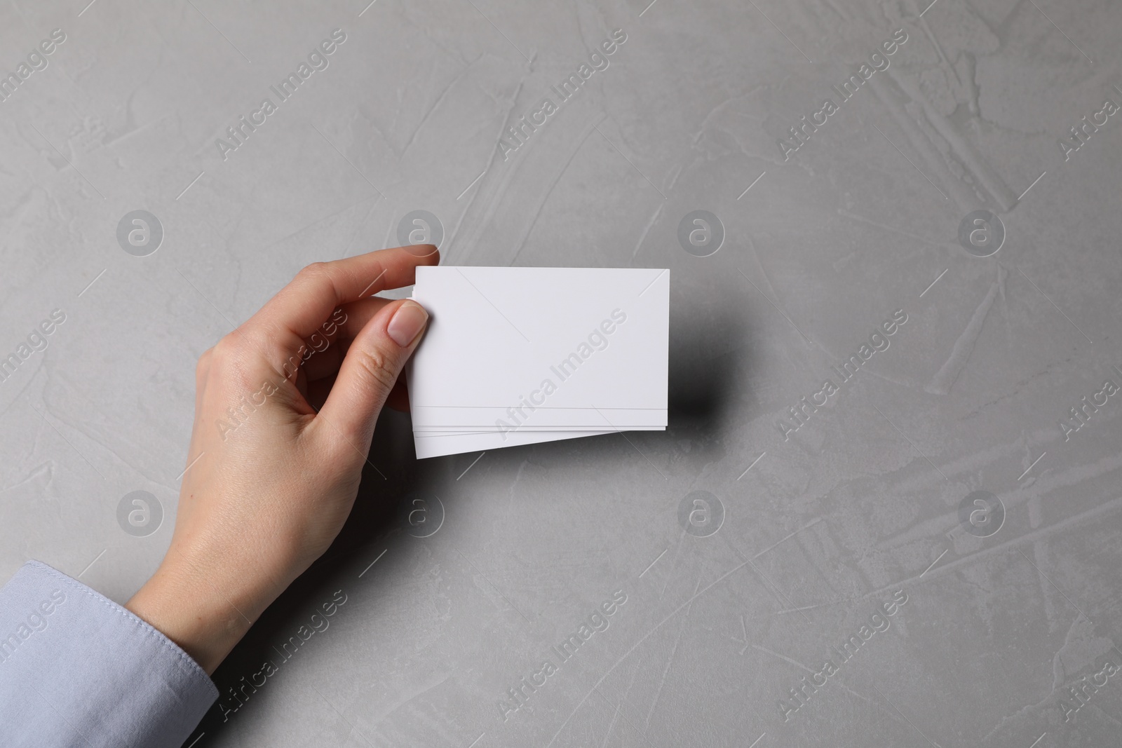 Photo of Woman holding blank cards at light grey table, top view. Mockup for design