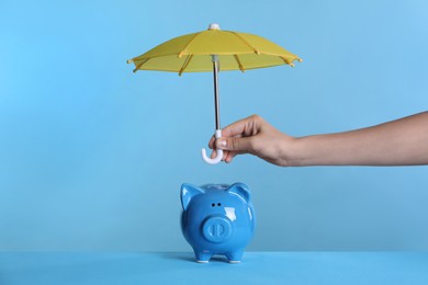 Woman holding small umbrella over piggy bank on light blue background, closeup