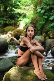 Photo of Beautiful young woman in stylish bikini relaxing on rock near mountain stream in forest