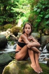 Beautiful young woman in stylish bikini relaxing on rock near mountain stream in forest