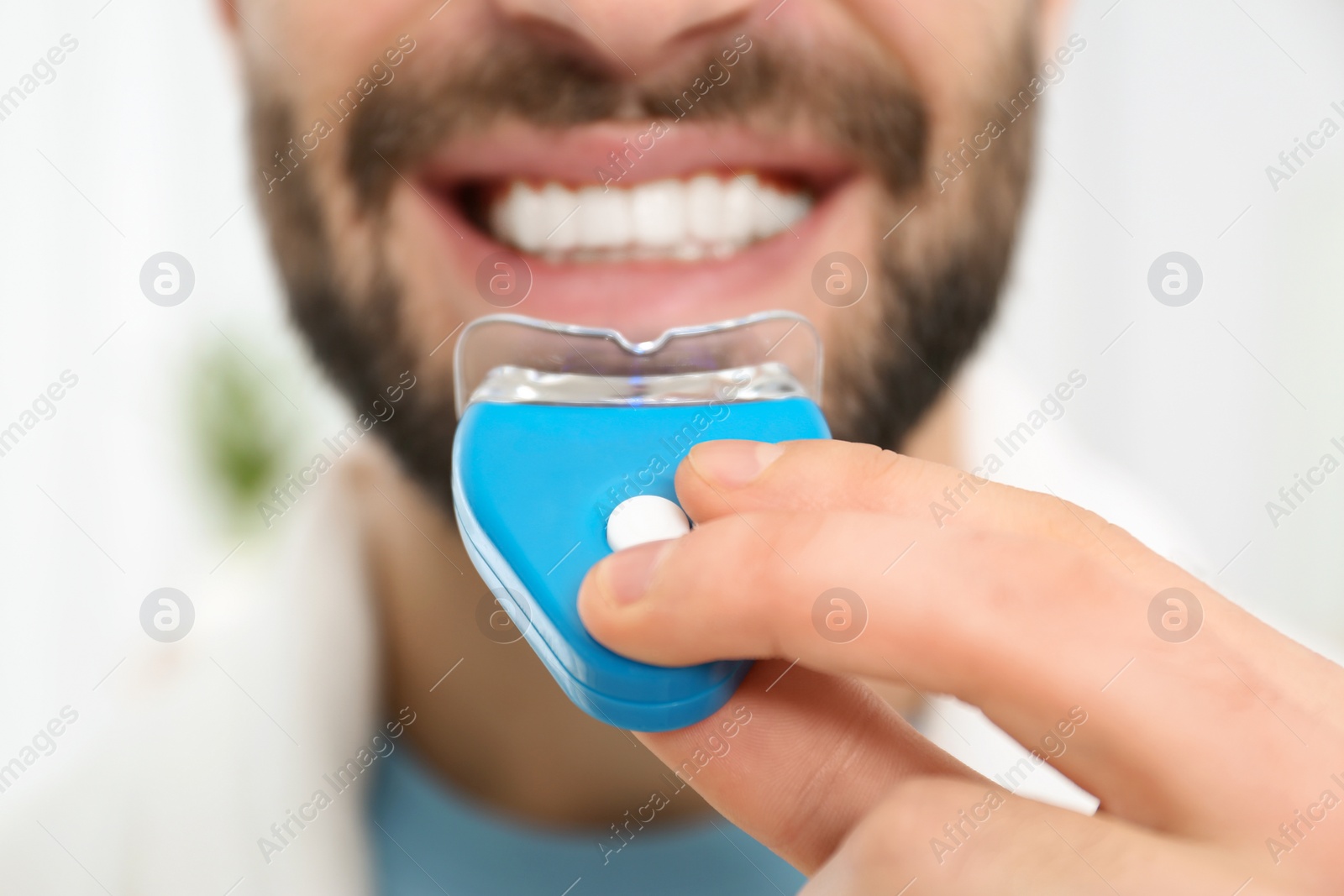 Photo of Young man using teeth whitening device on light background, closeup