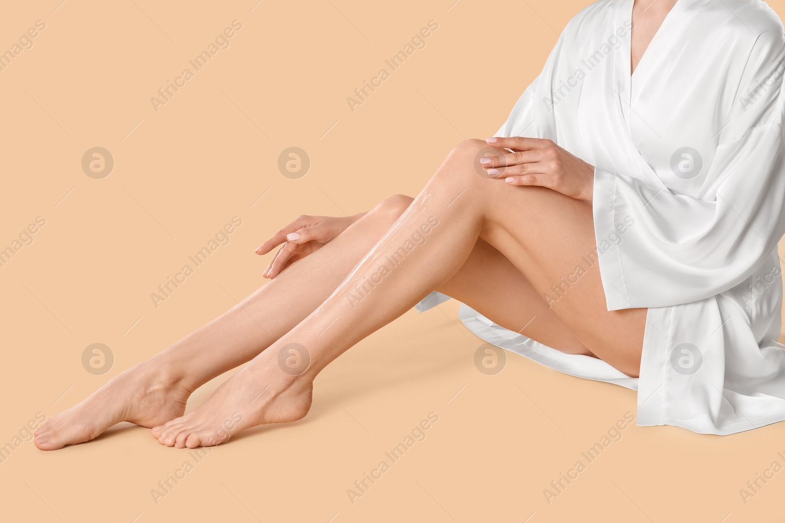 Photo of Woman applying body cream onto her smooth legs on beige background, closeup