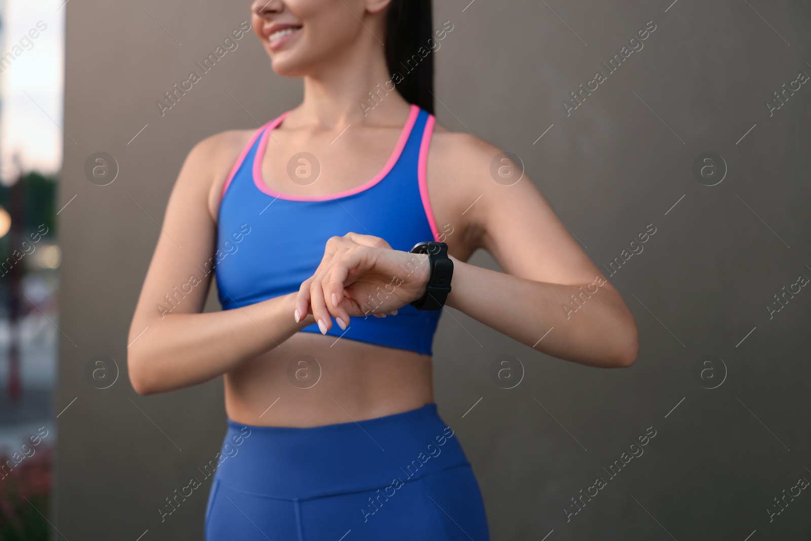 Photo of Woman in stylish sportswear checking smart watch near black wall outdoors, closeup