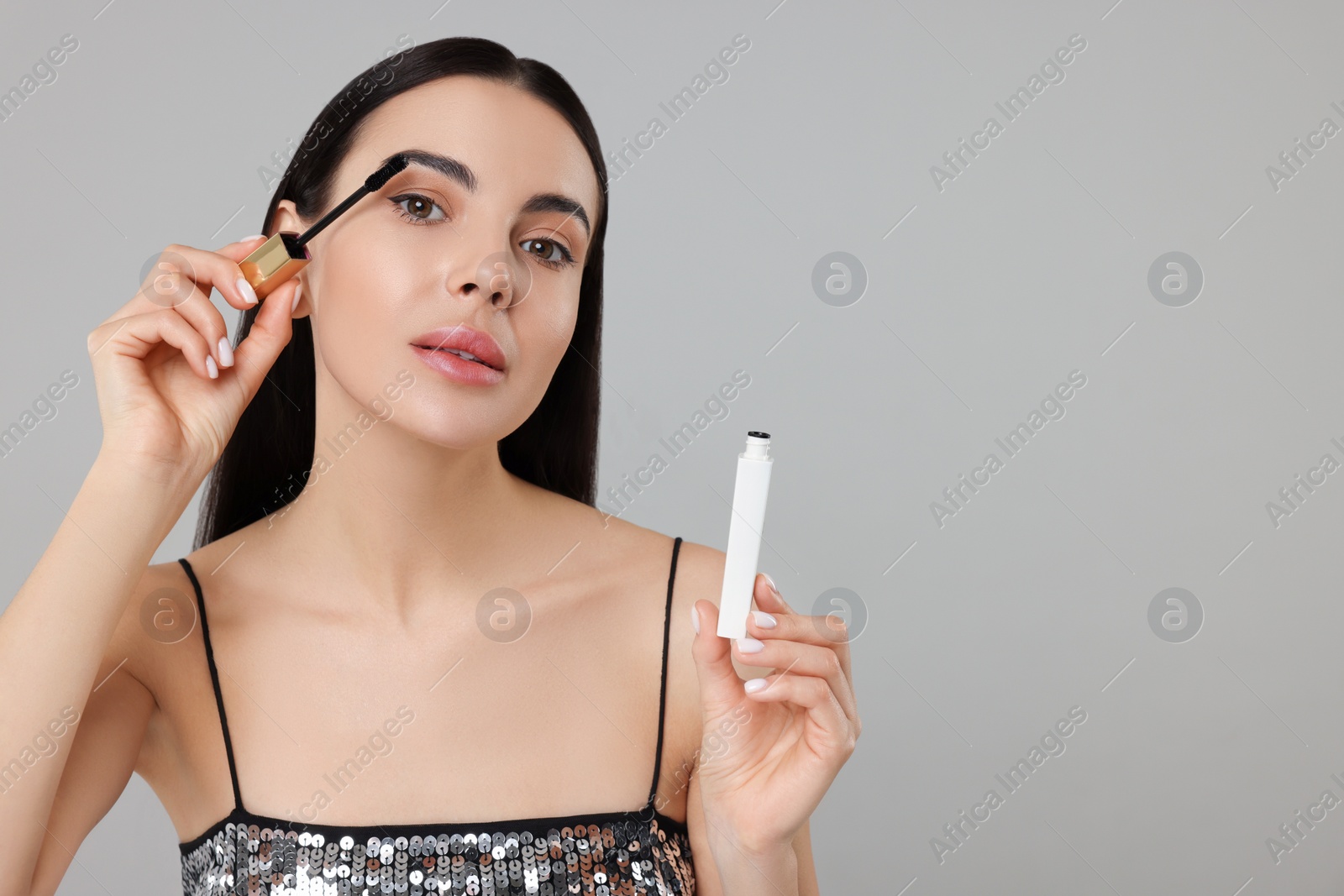 Photo of Beautiful young woman applying mascara on grey background, space for text