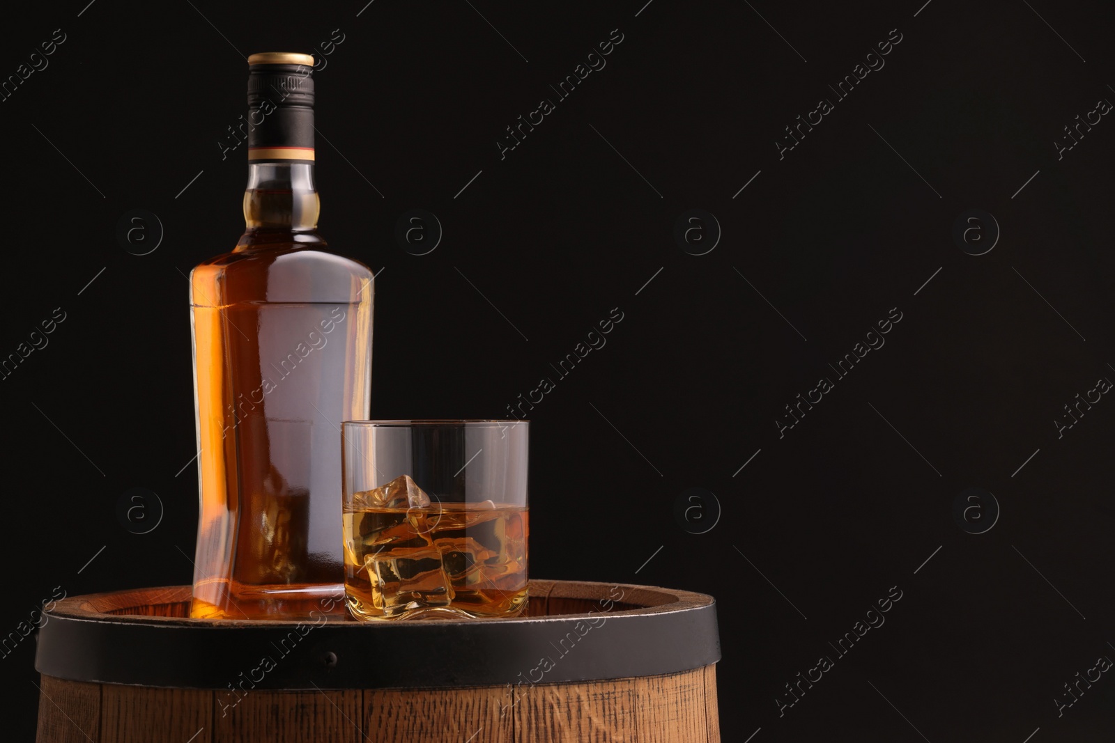 Photo of Whiskey with ice cubes in glass and bottle on wooden barrel against black background, space for text