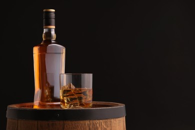 Whiskey with ice cubes in glass and bottle on wooden barrel against black background, space for text