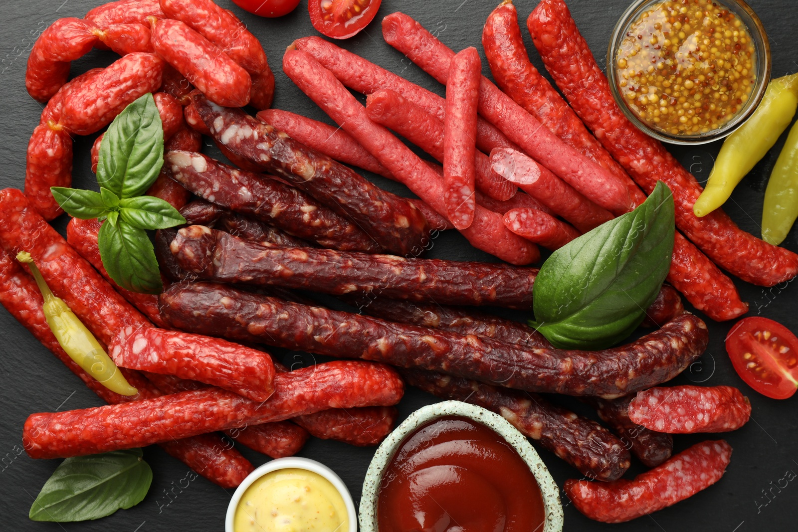 Photo of Different thin dry smoked sausages, basil and sauces on black table, flat lay