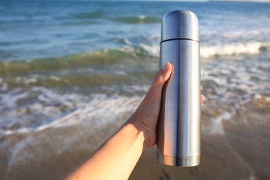 Woman holding metallic thermos with hot drink on beach near sea, closeup