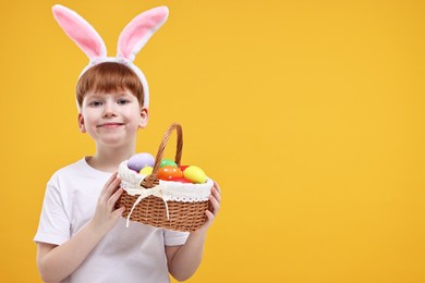 Easter celebration. Cute little boy with bunny ears and wicker basket full of painted eggs on orange background. Space for text